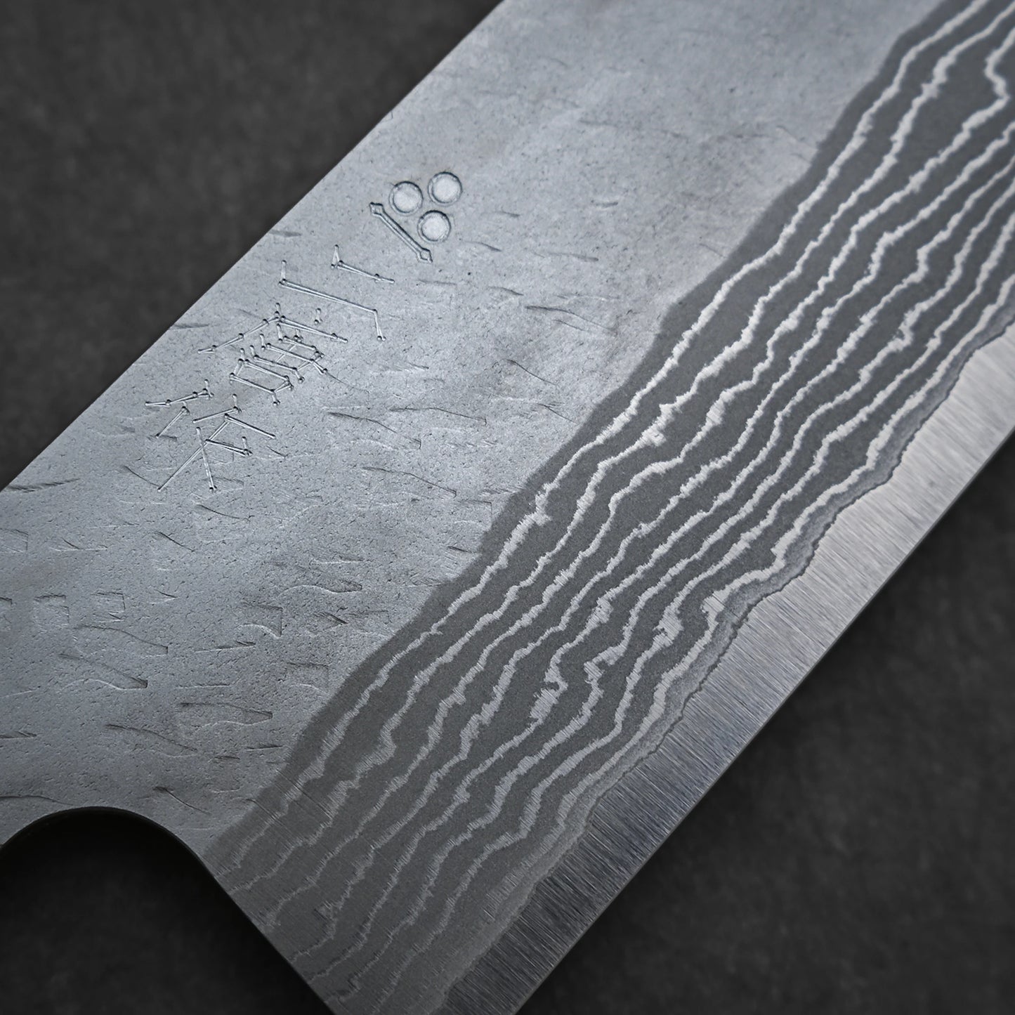Close-up of the Nigara Hamono Nigara tsuchime damascus VG10 gyuto 210mm blade, showcasing its intricate wavy patterns and etched markings on the metal surface against a dark background.