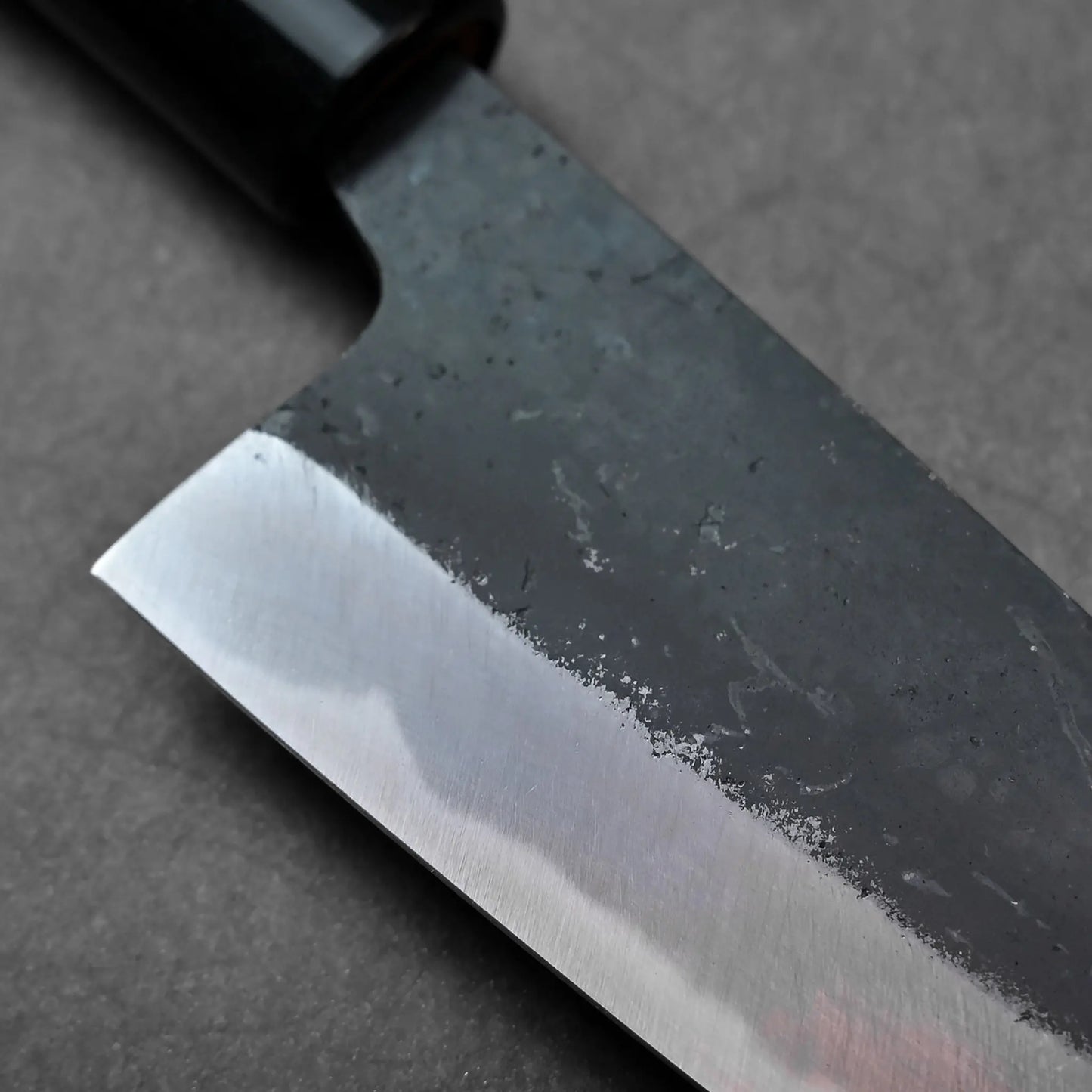 Close-up shows the blade of a Japanese knife, with a textured, dark upper section and part of its slender shape visible.