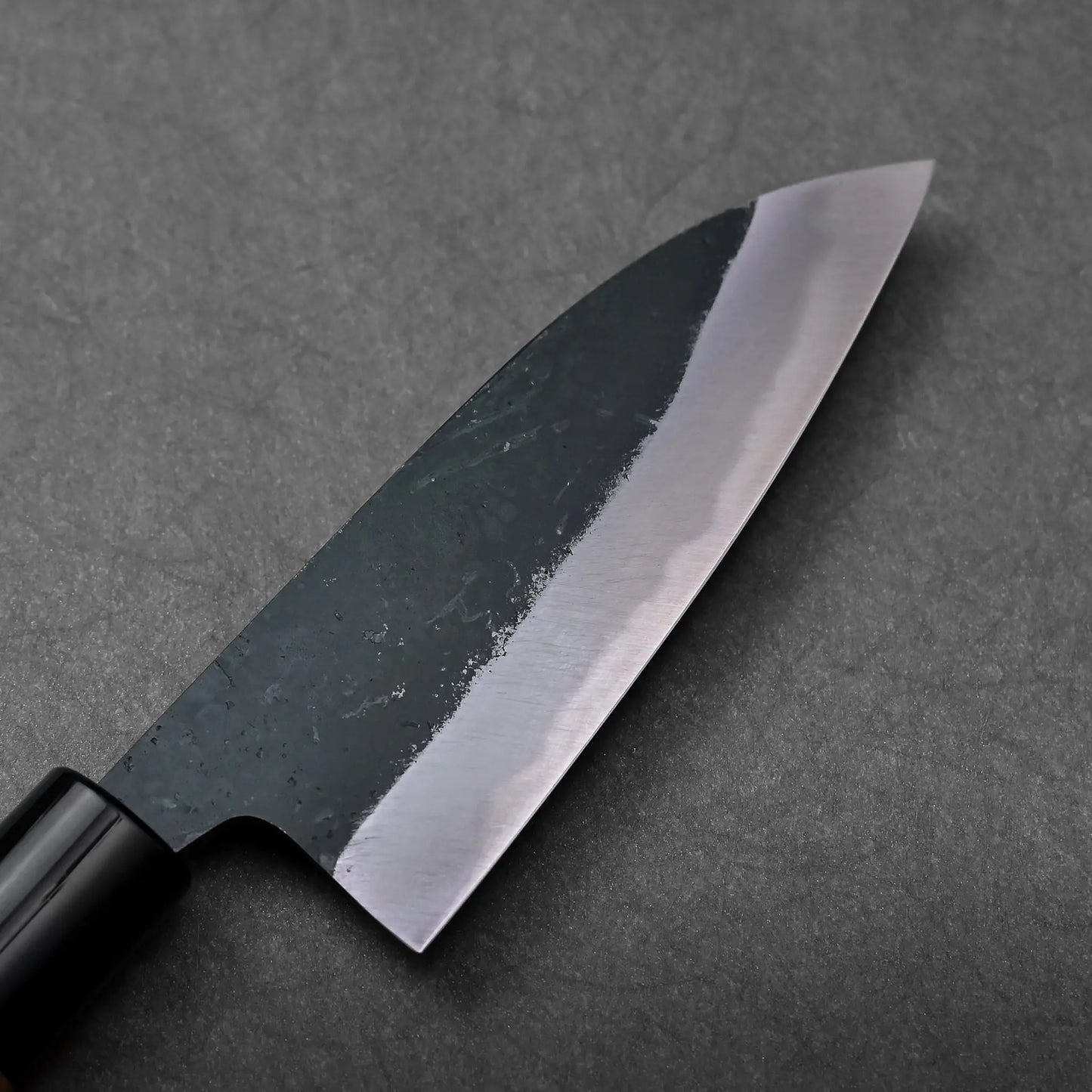 A close-up showing the blade of a Japanese knife, with hand-forged texture and a black and silver finish.