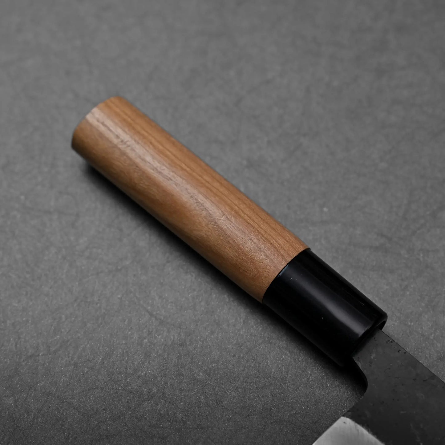 A close-up of a wooden handle with black bolster on a Japanese knife, blade details not visible in this image.