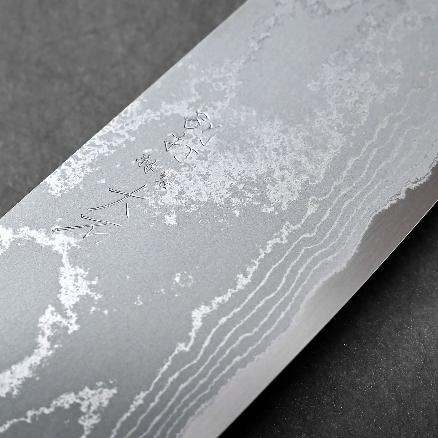 Blade close-up showing damascus cladding, engraved characters visible on Japanese knife metal, santoku shape implied.