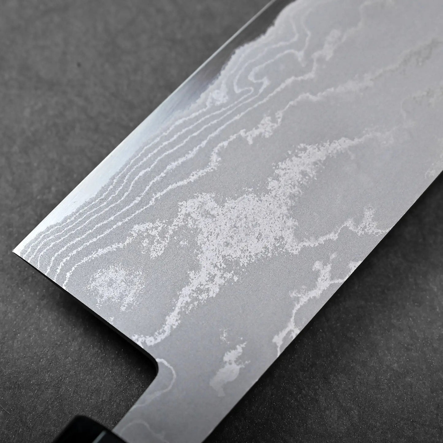 Blade close-up showing a Japanese knife with a straight edge and rounded tip, featuring wavy Damascus patterns.