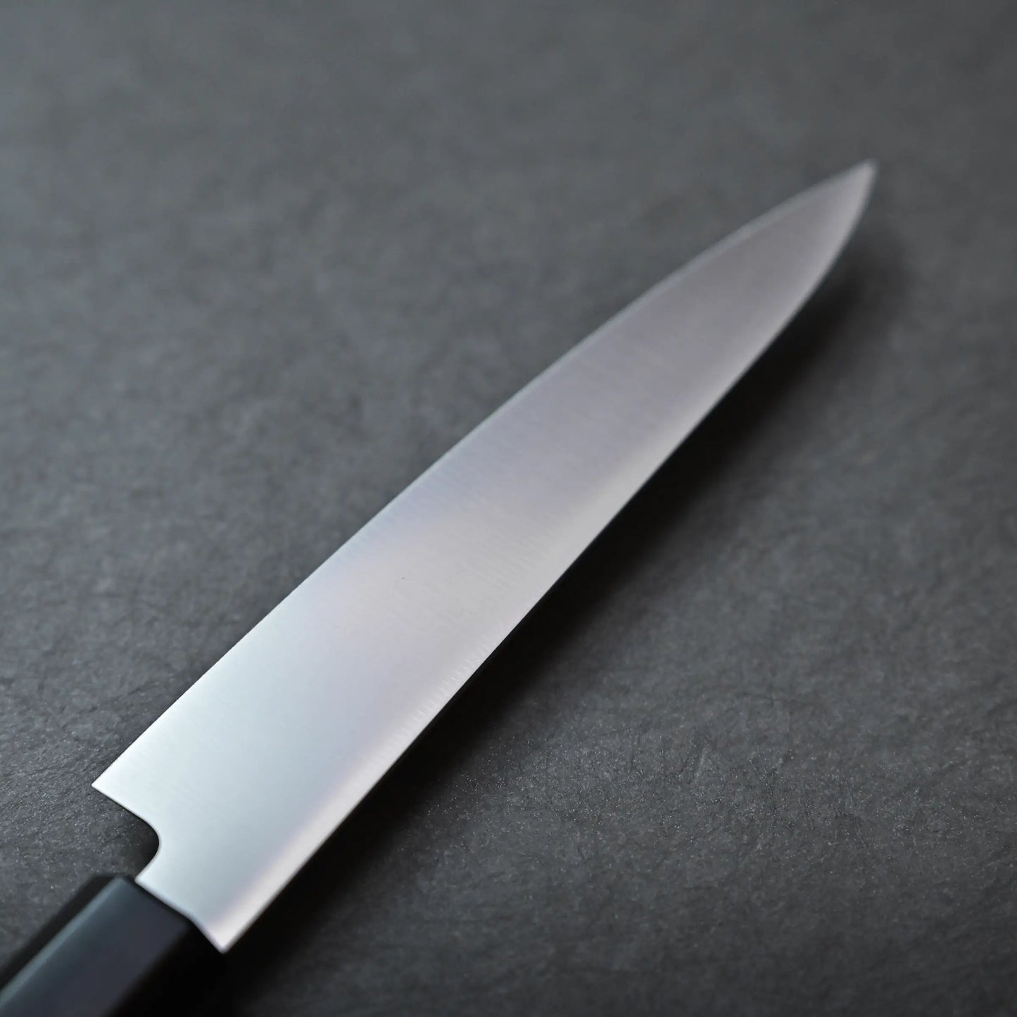 A close-up view of a stainless petty knife with a black handle, resting on a dark gray textured surface.