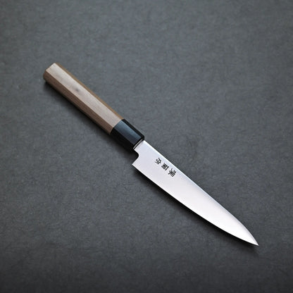 A Japanese-style Sakai petty knife with a wooden handle and a pointed stainless steel blade features Japanese characters engraved near the handle. The knife rests on a dark, textured surface.