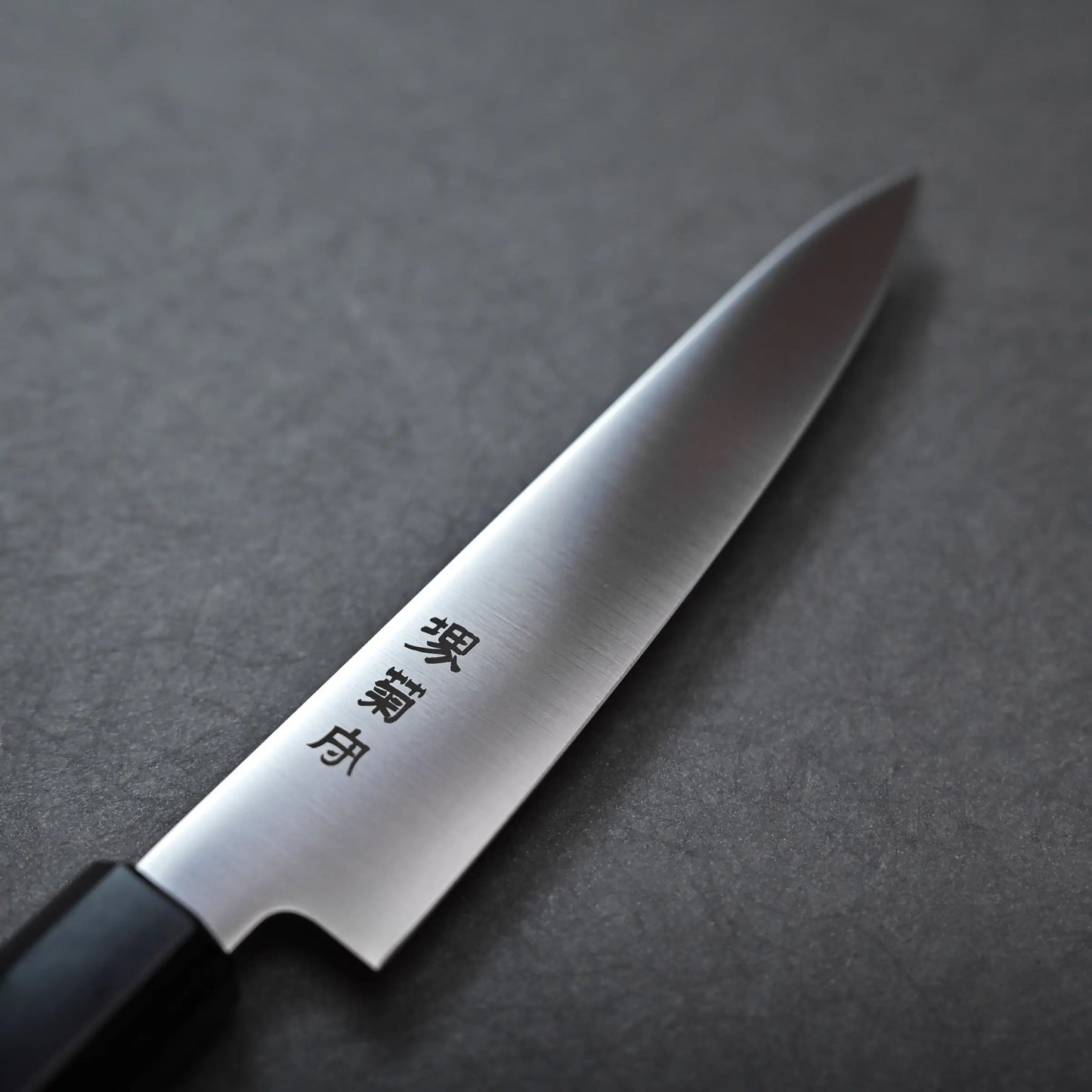 A close-up of a Sakai petty knife with a black handle and Japanese characters engraved on the stainless steel blade, resting on a dark textured surface.