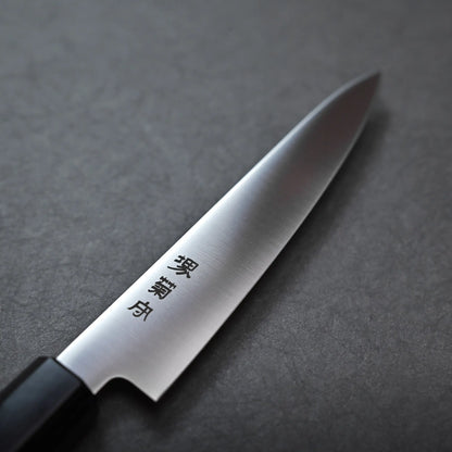 A close-up of a Sakai petty knife with a black handle and Japanese characters engraved on the stainless steel blade, resting on a dark textured surface.