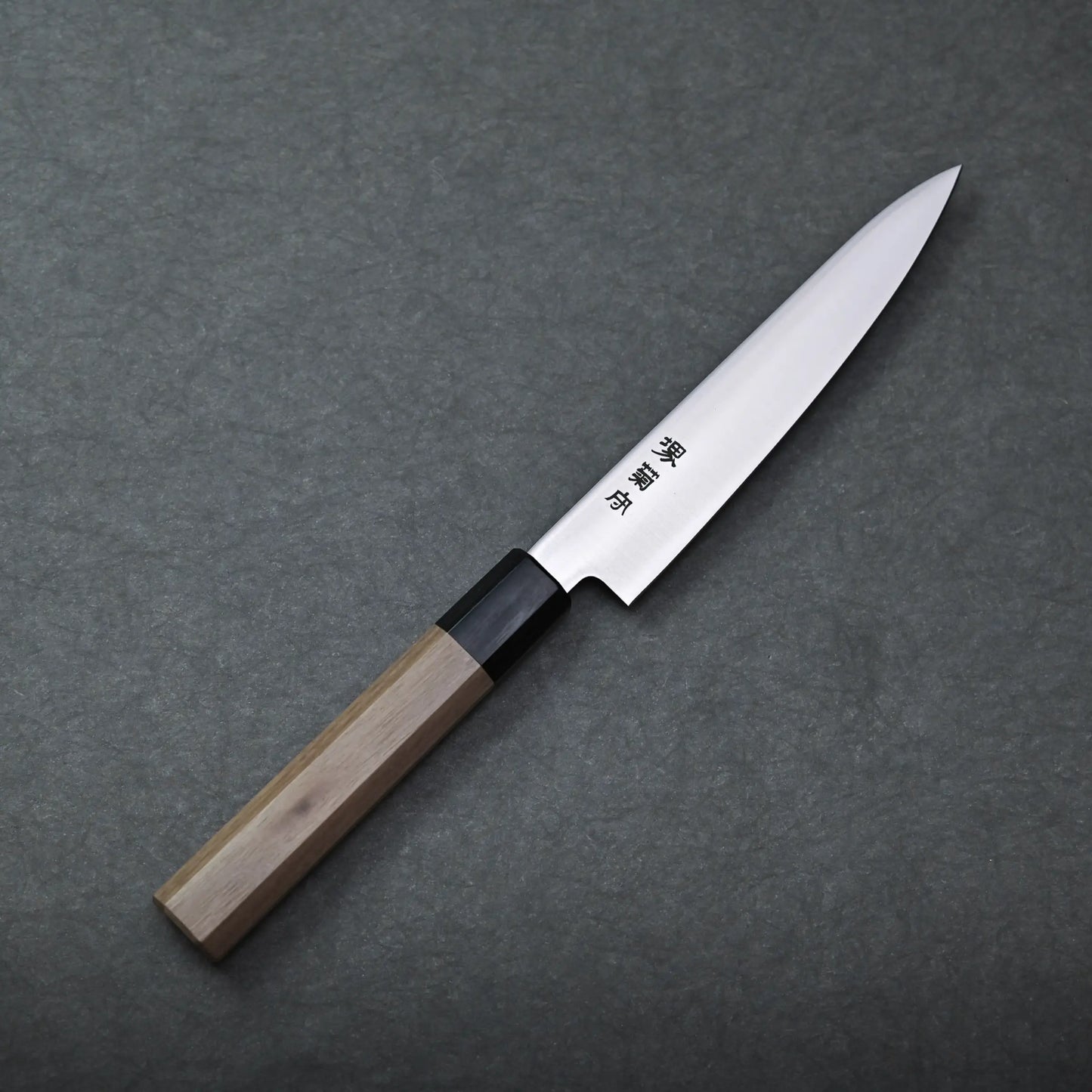 A Sakai petty knife with a pointed stainless blade and a wooden handle rests on a dark gray textured surface. The blade displays engraved Japanese characters.