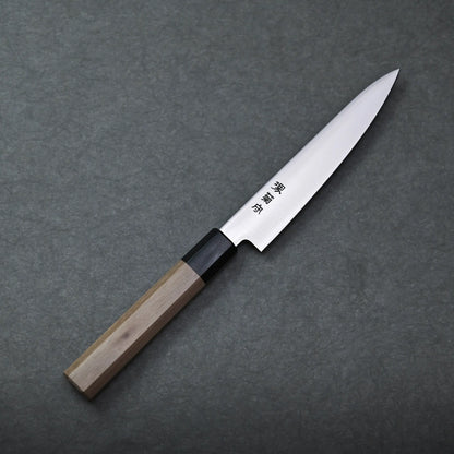 A Sakai petty knife with a pointed stainless blade and a wooden handle rests on a dark gray textured surface. The blade displays engraved Japanese characters.