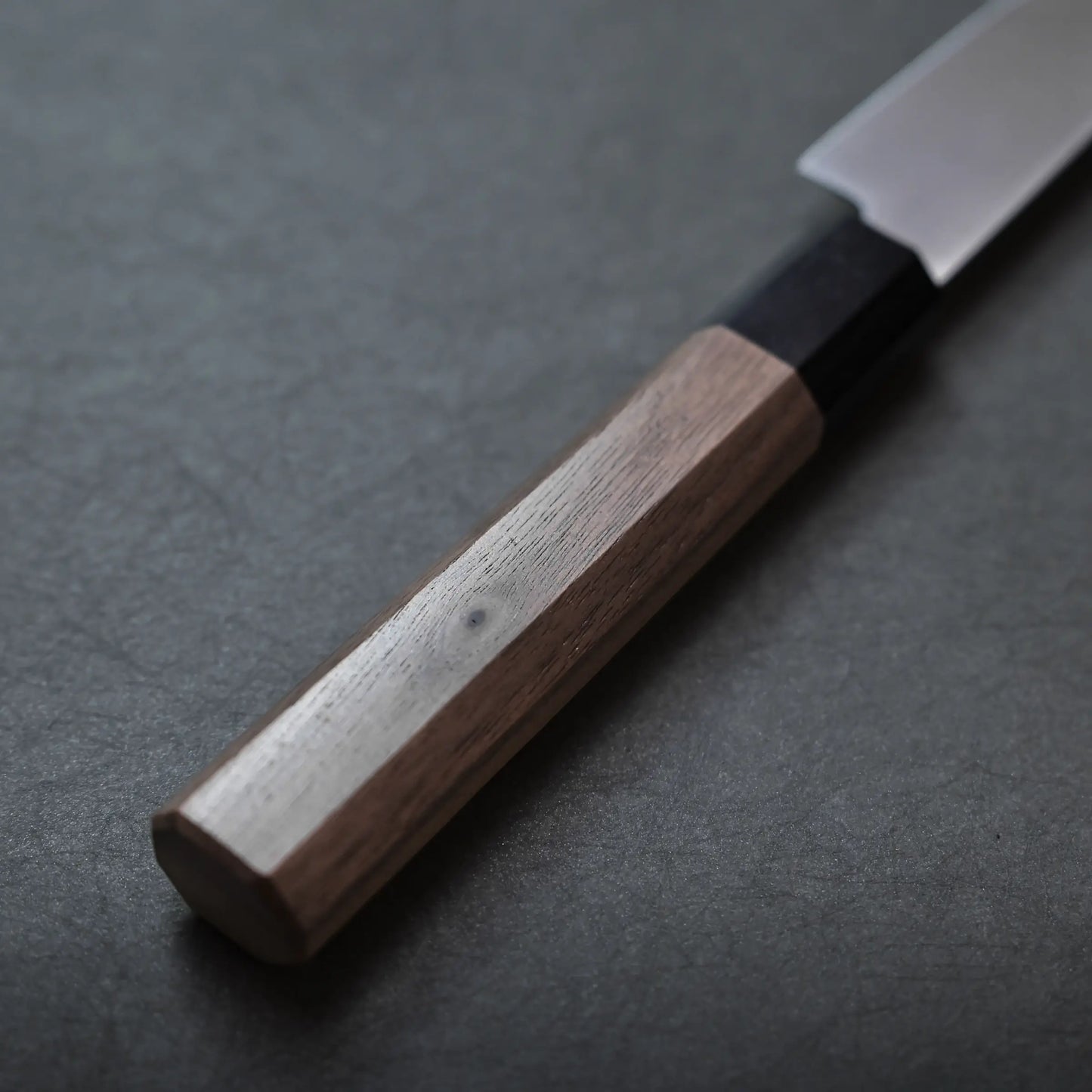 A close-up of a Sakai petty knife with a wooden handle and stainless blade, positioned on a dark gray textured surface.