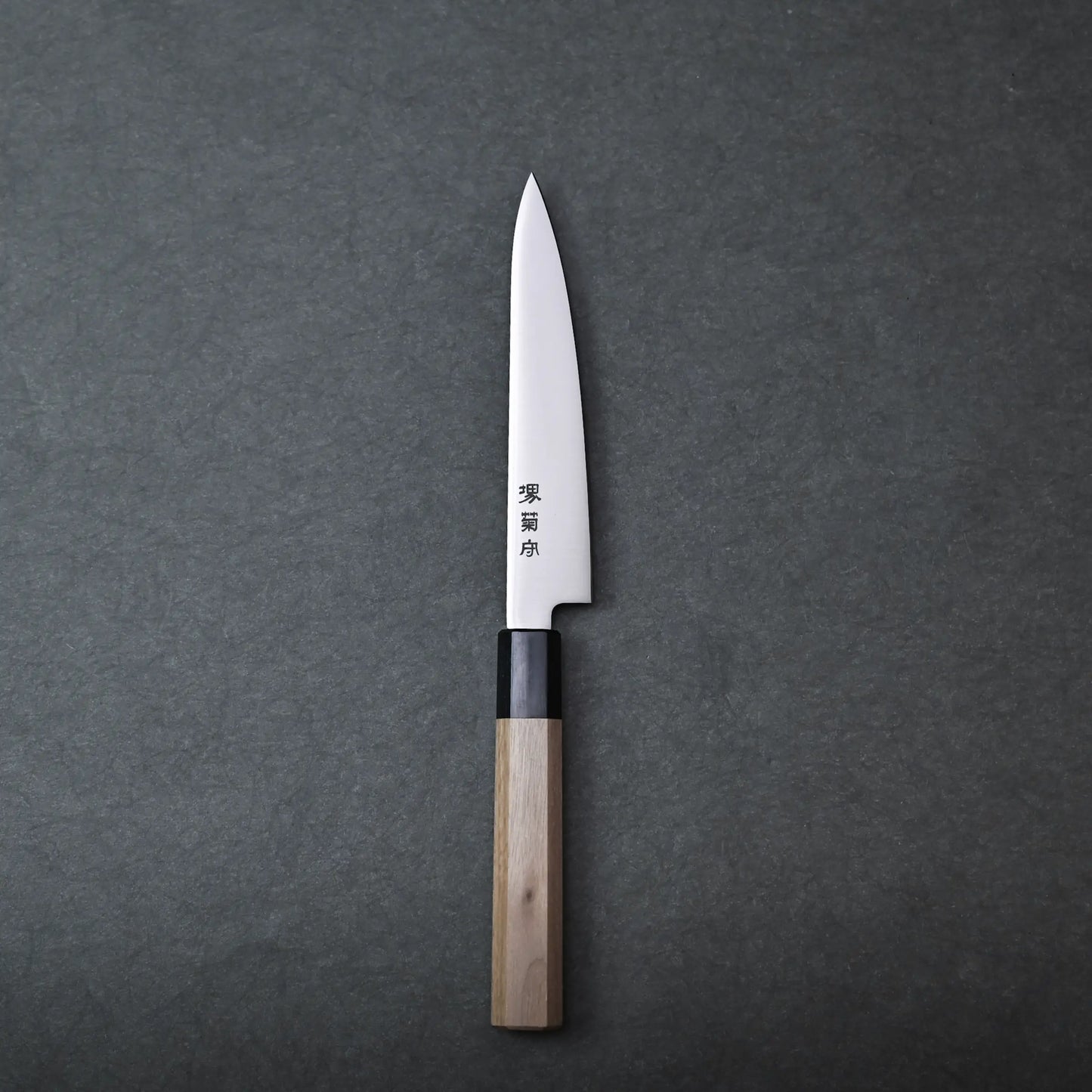 A single Sakai petty knife with a wooden handle and a pointed blade rests on a dark gray surface. Japanese characters are engraved on the stainless blade.