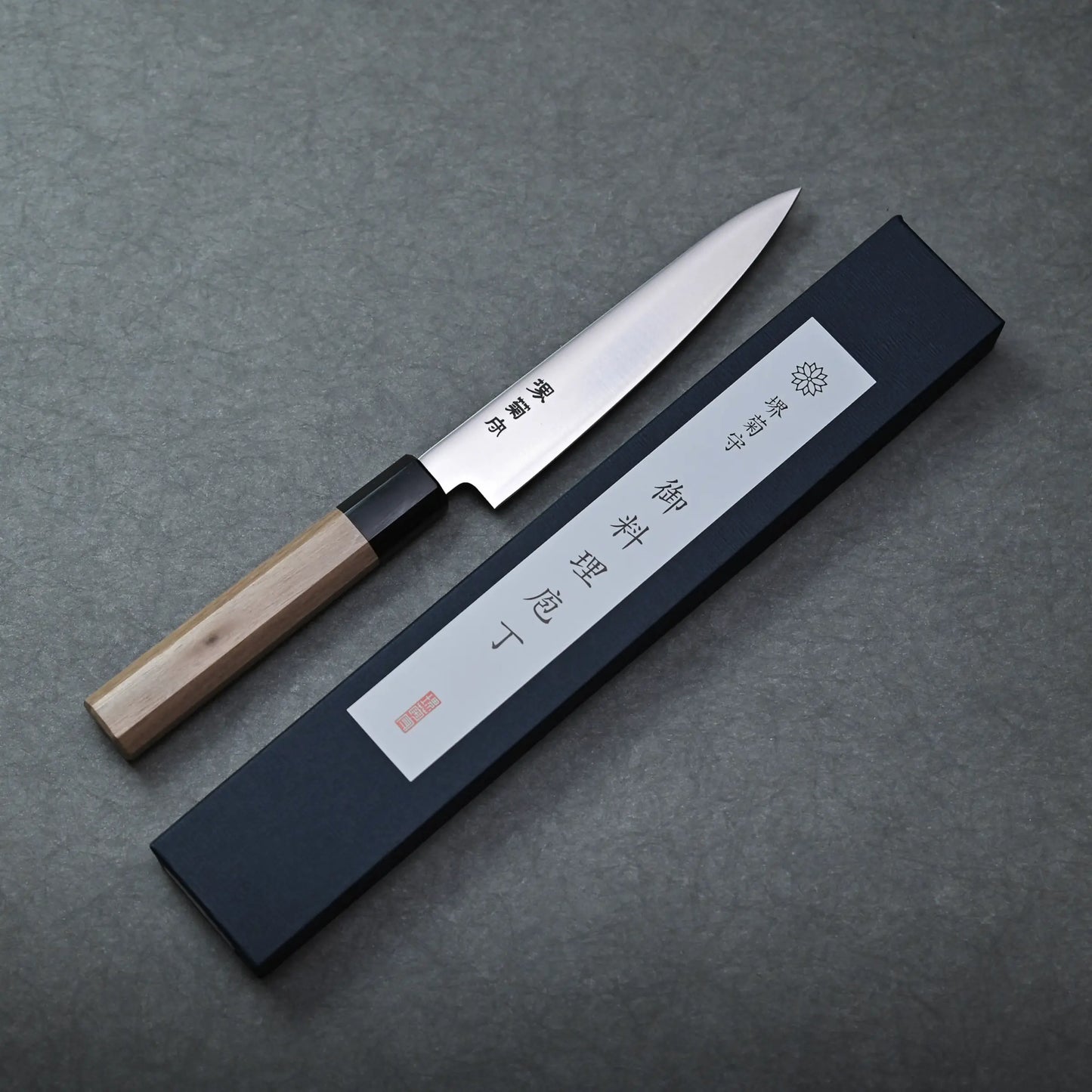 A Sakai petty knife with a wooden handle rests beside a black rectangular box featuring Japanese characters on a gray surface.