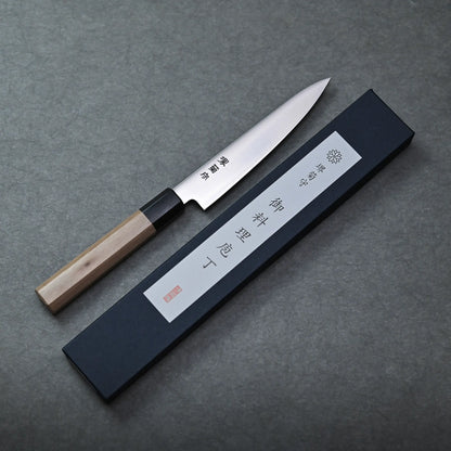 A Sakai petty knife with a wooden handle rests beside a black rectangular box featuring Japanese characters on a gray surface.