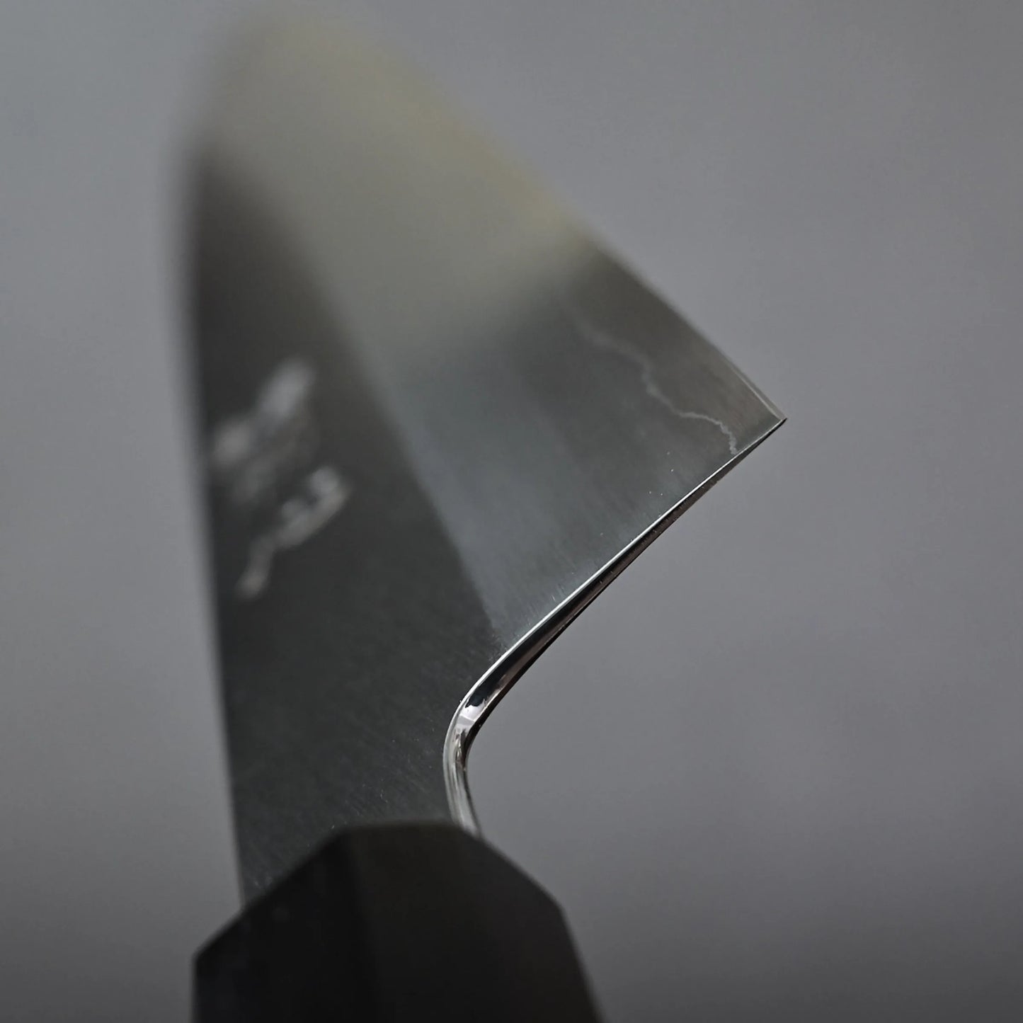 Close-up of the sharp edge and tip of a gyuto knife with a black handle, set against a plain grey background. The blade shows a subtle wavy pattern near the edge.