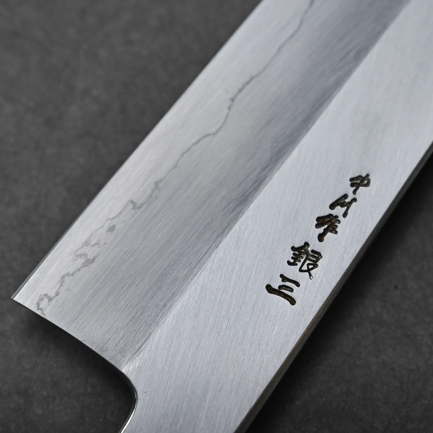 Close-up of a gyuto knife blade with a visible wavy pattern near the edge and black Japanese characters engraved on the flat surface. The background is a dark, plain surface.