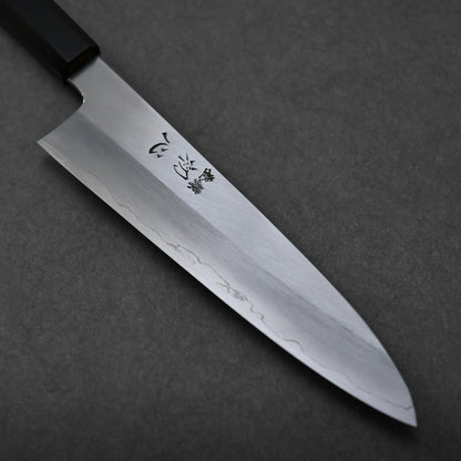 A close-up of a gyuto knife with a sharp, shiny blade featuring engraved Japanese characters and a distinct wavy pattern near the edge, set on a dark textured surface.