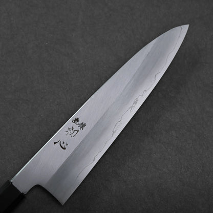 A close-up of a gyuto knife, a Japanese chef’s knife with a shiny steel blade engraved with Japanese characters, lying on a dark textured surface. The blade shows a visible temper line.