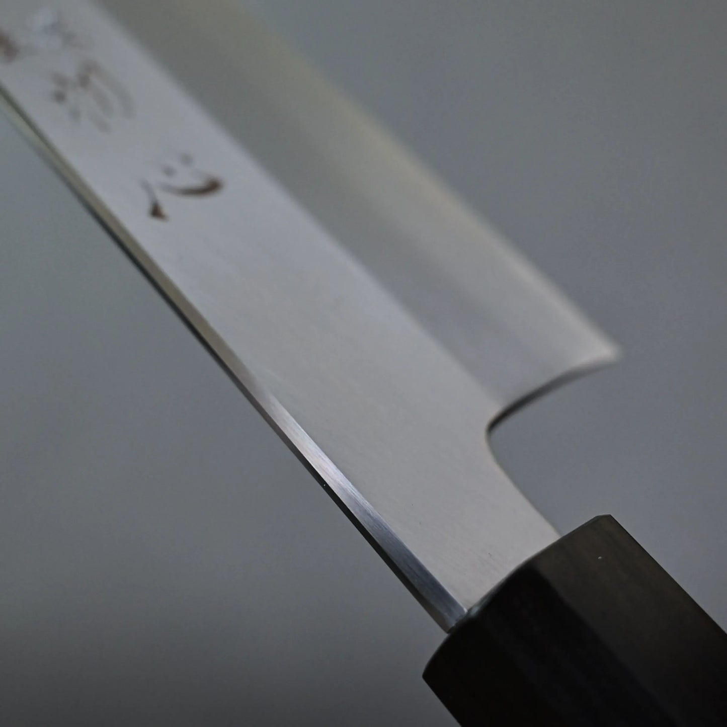 Close-up of a gyuto knife blade, showing a polished metal surface with engraved characters and a dark handle, set against a plain gray background.