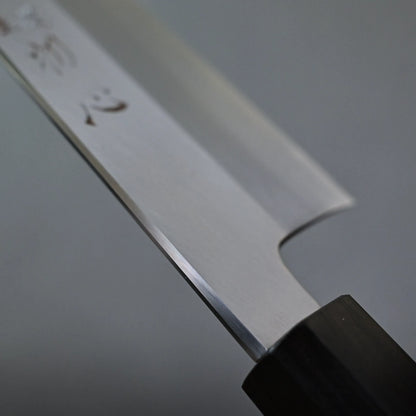 Close-up of a gyuto knife blade, showing a polished metal surface with engraved characters and a dark handle, set against a plain gray background.