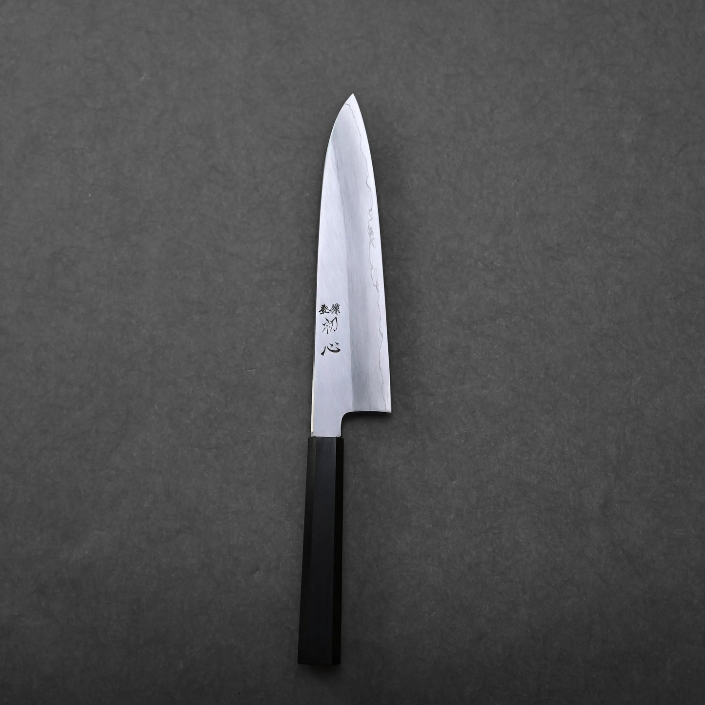 A Japanese chef's knife with a black rectangular handle and a shiny silver blade featuring engraved characters, photographed on a dark gray background. This gyuto knife embodies traditional craftsmanship and modern elegance.
