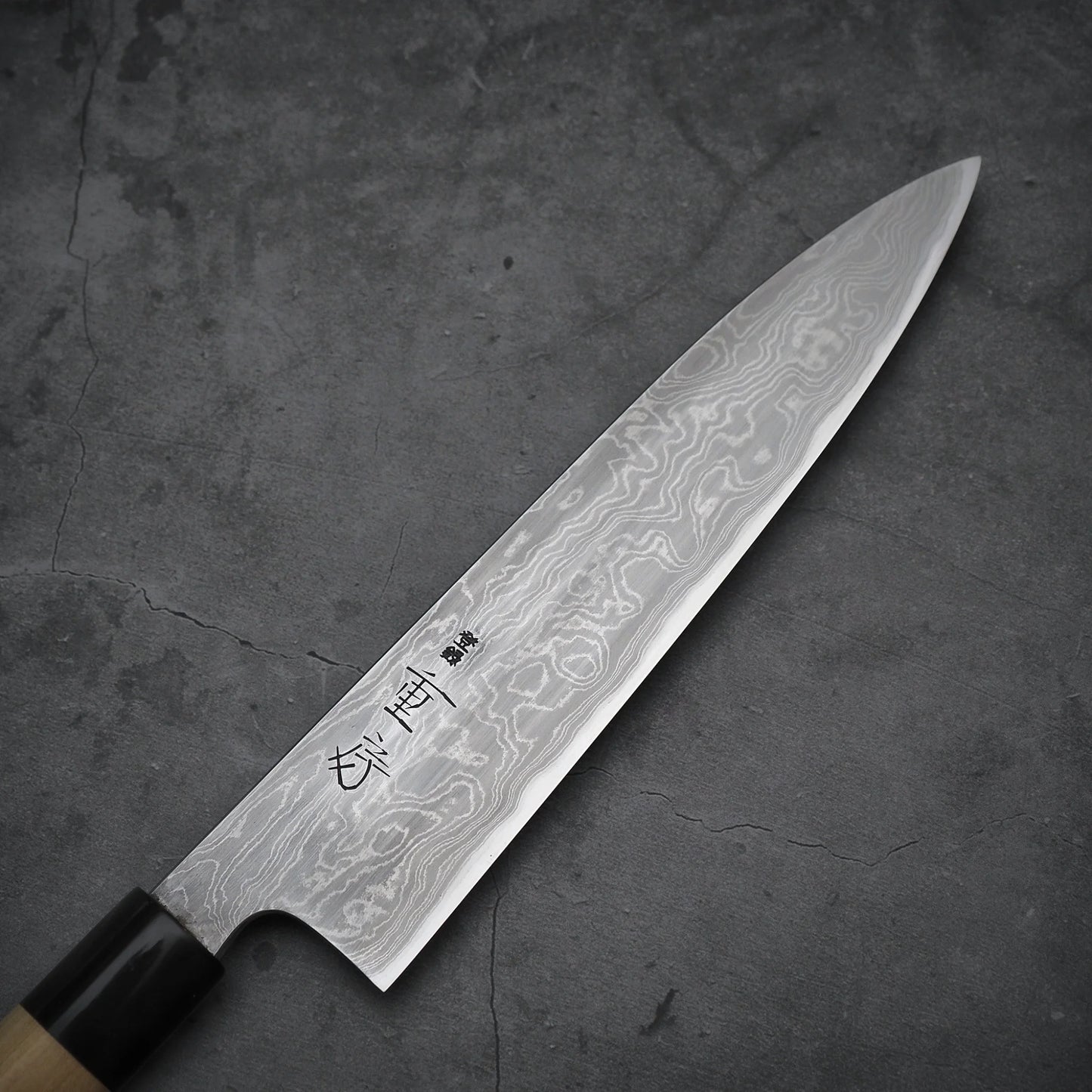 Top-down view of a 210mm gyuto with kitaeji finish and light wood handle, placed on a dark surface. Shigefusa kitaeji model.