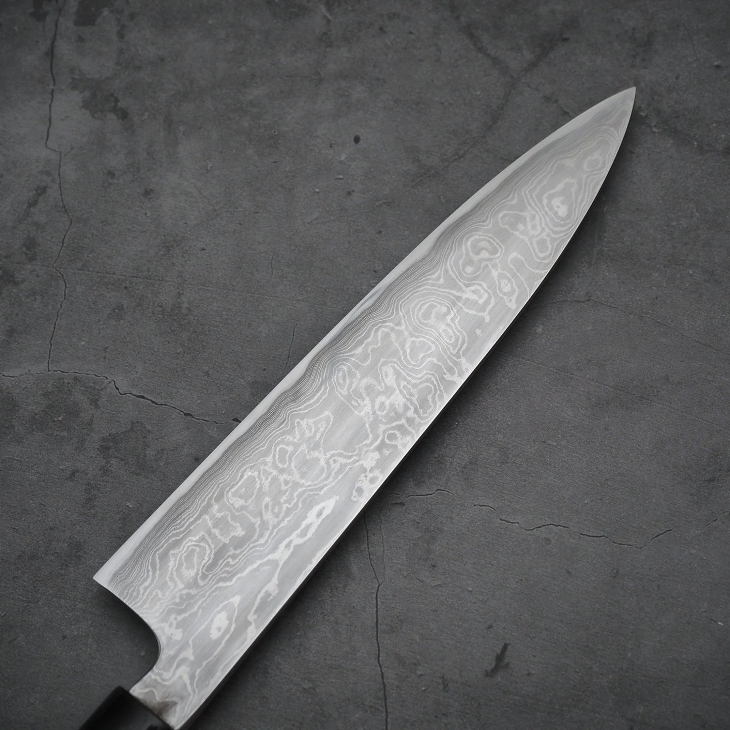 Close-up view of the reverse side of a damascus blade, showing wavy steel patterns and a clean surface. Background is a textured gray slab.