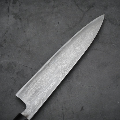 Close-up view of the reverse side of a damascus blade, showing wavy steel patterns and a clean surface. Background is a textured gray slab.