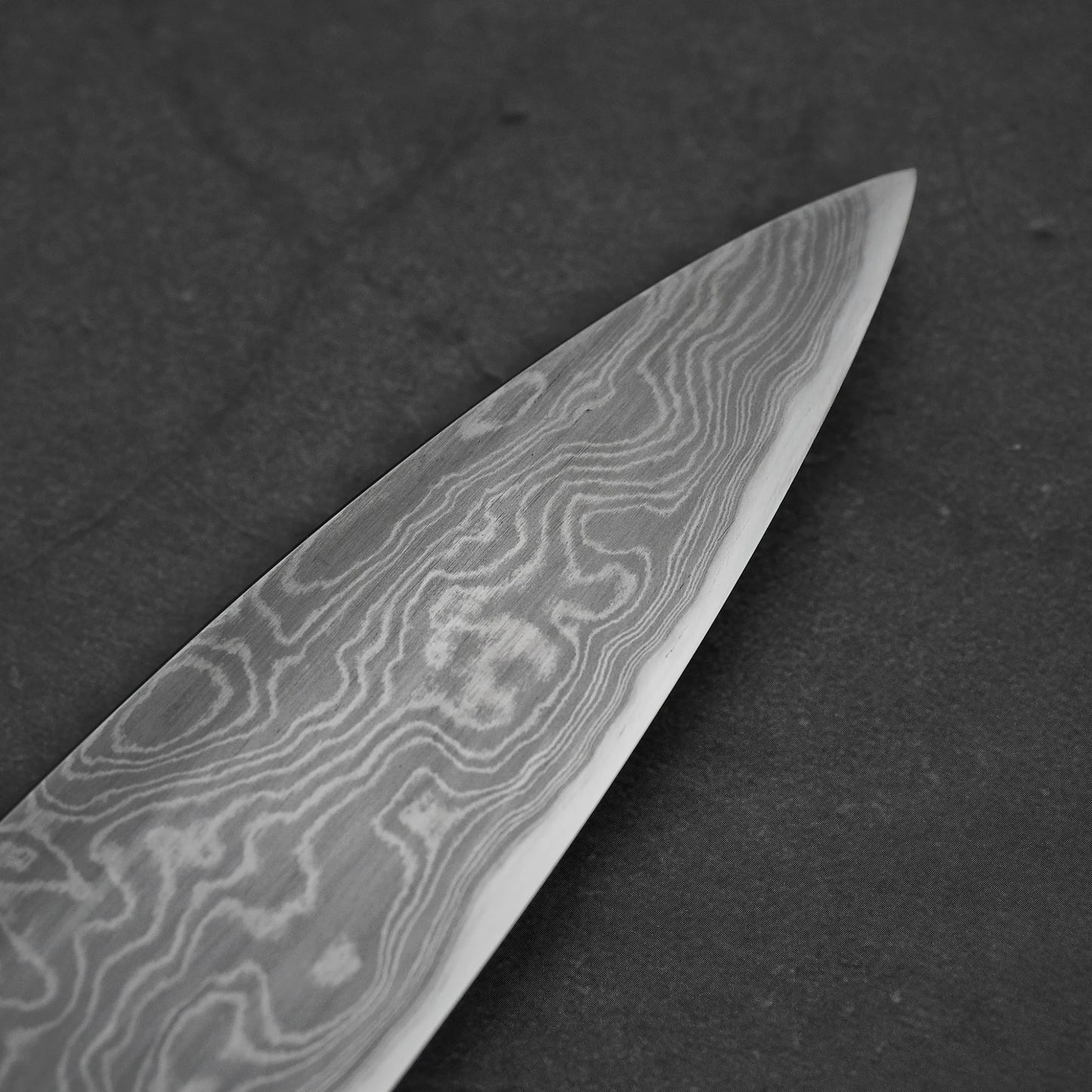 Close-up of a damascus steel knife blade on a dark background showing the tip area