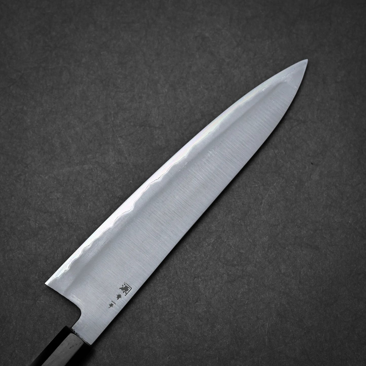 A close-up of a kasumi Japanese chef's knife with a sharp, pointed blade and a dark handle, set against a dark gray textured background.