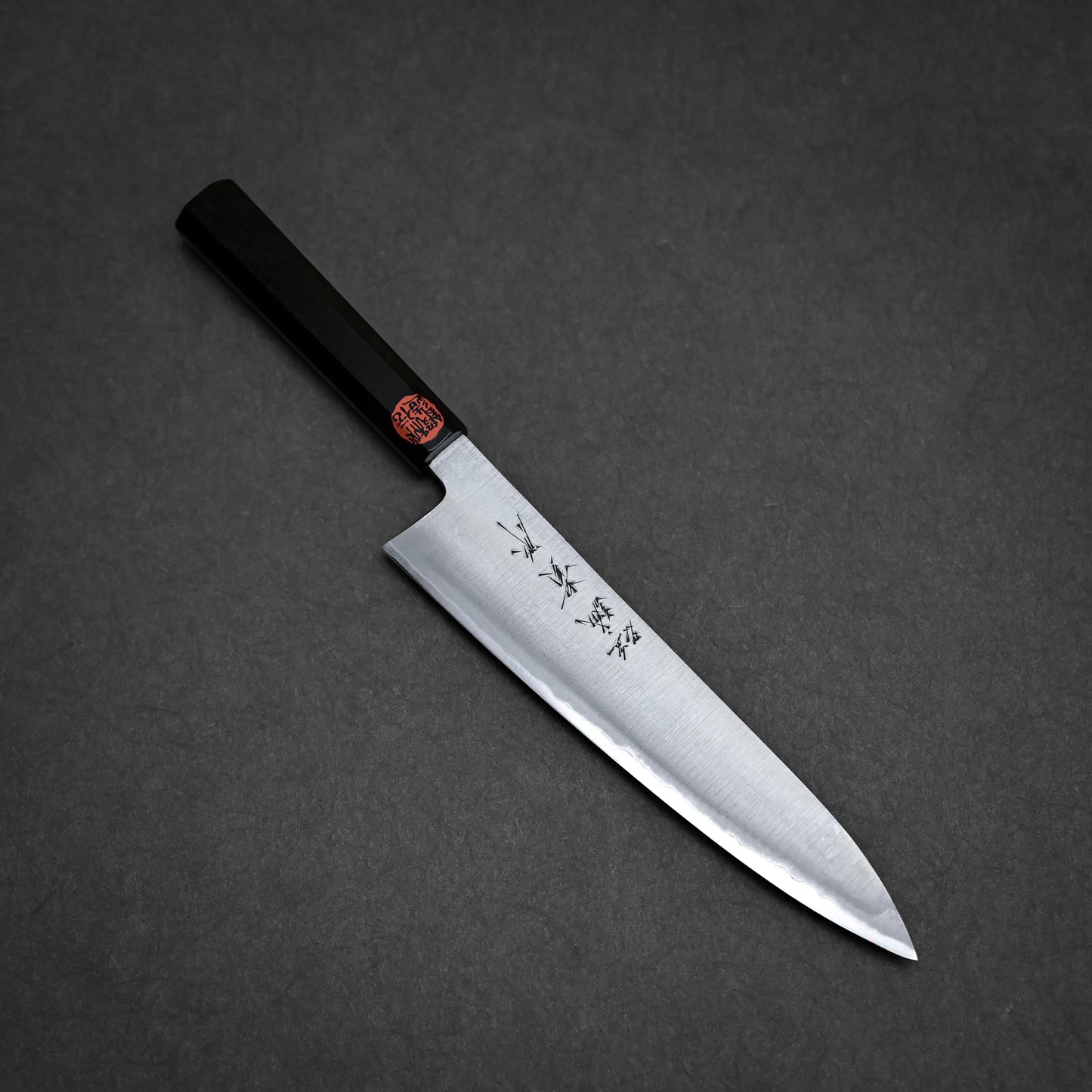 A kasumi chef's knife with a dark handle and a steel blade featuring engraved Japanese characters, shown on a dark gray textured surface.