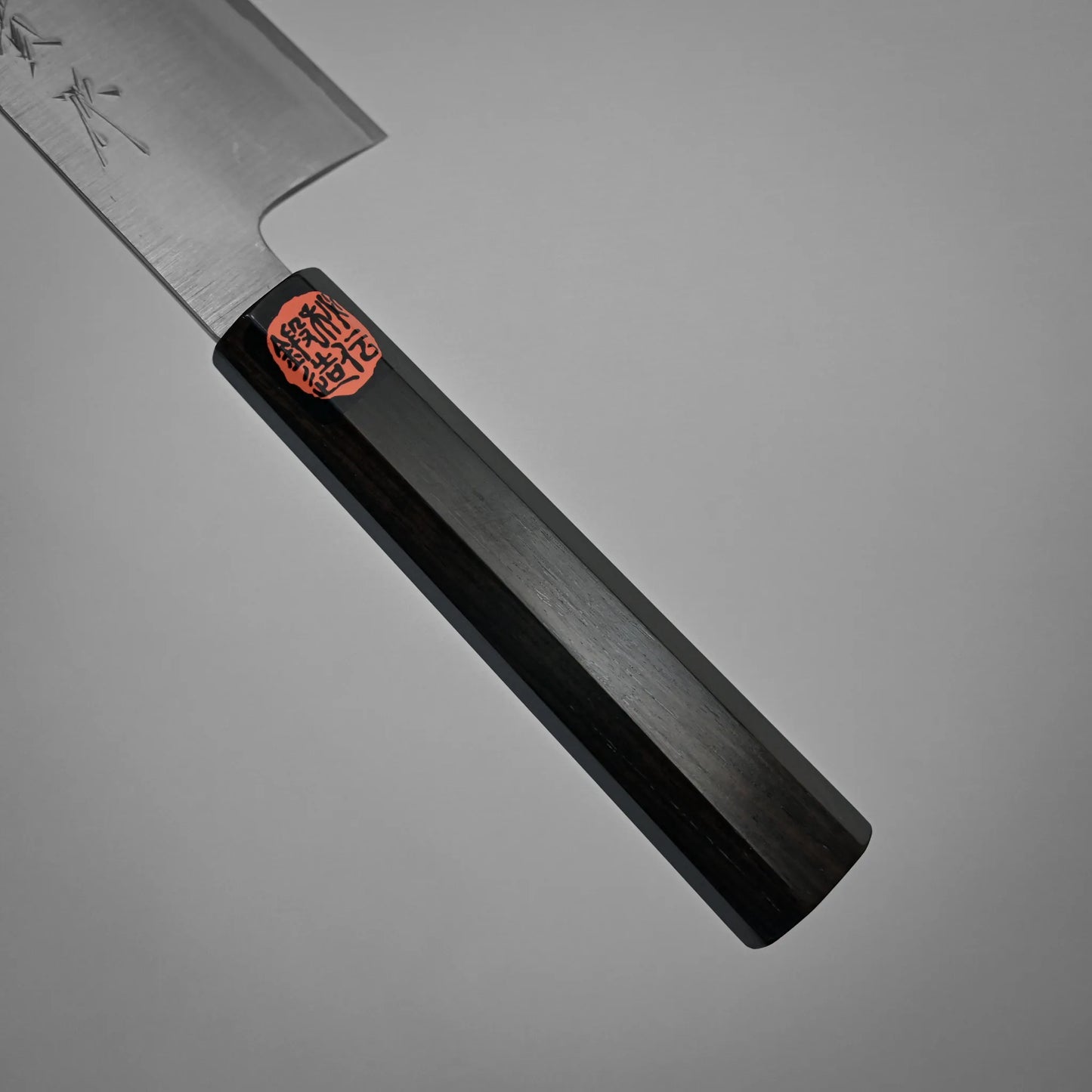 Close-up of a kasumi chef's knife handle made of dark wood with a red and black sticker featuring Japanese characters, set against a plain gray background.