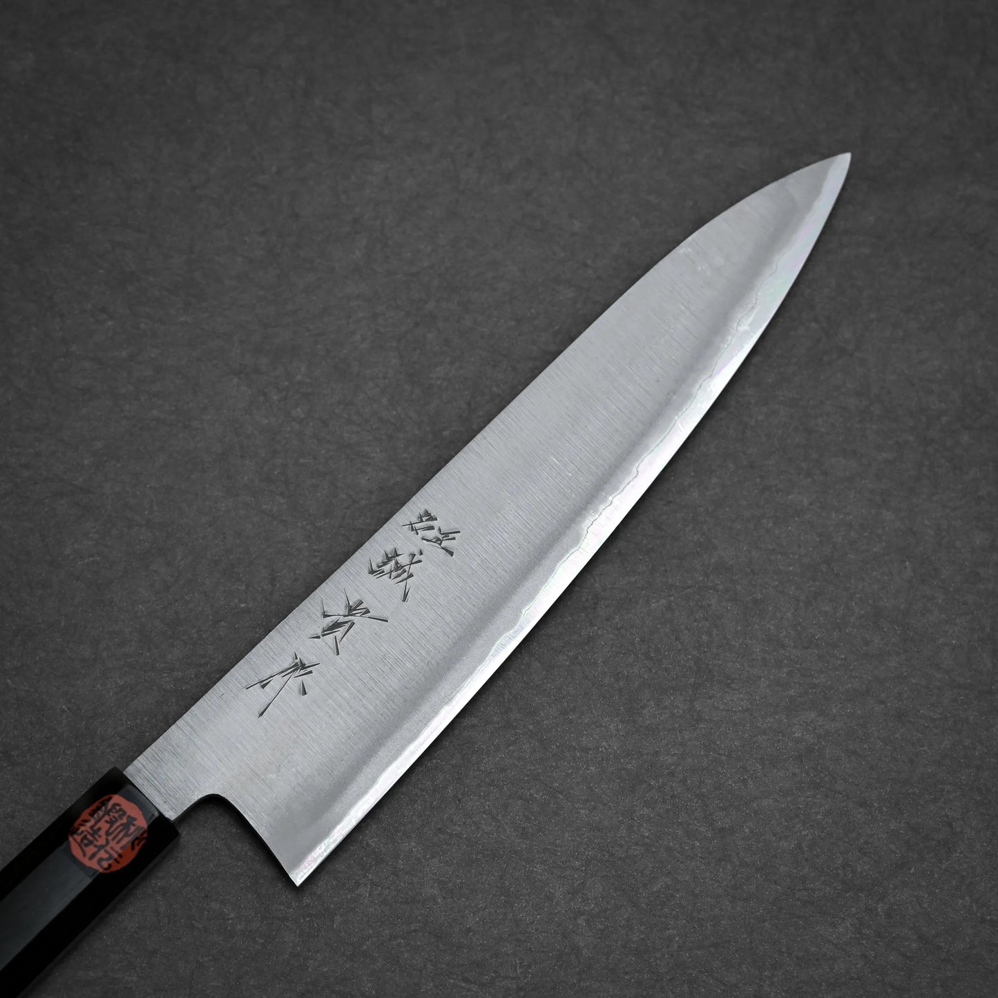 A kasumi gyuto Japanese-style kitchen knife with a sharp, pointed blade and kanji characters engraved on it, photographed on a dark gray textured surface.