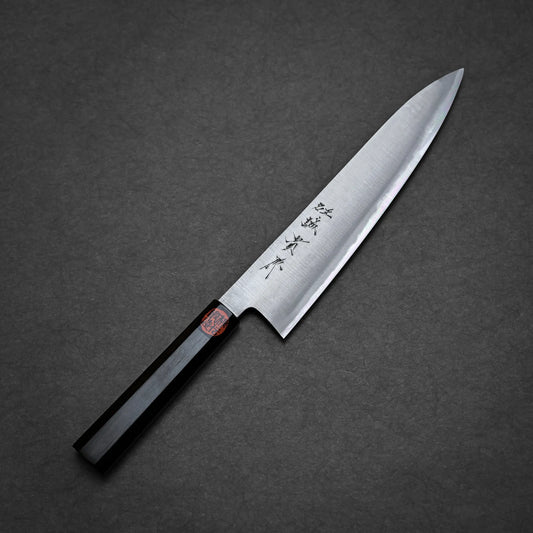 A Shigeki Tanaka migaki aogami#2 gyuto 240mm with a black handle and a silver blade featuring Japanese characters, placed diagonally on a dark gray textured surface.