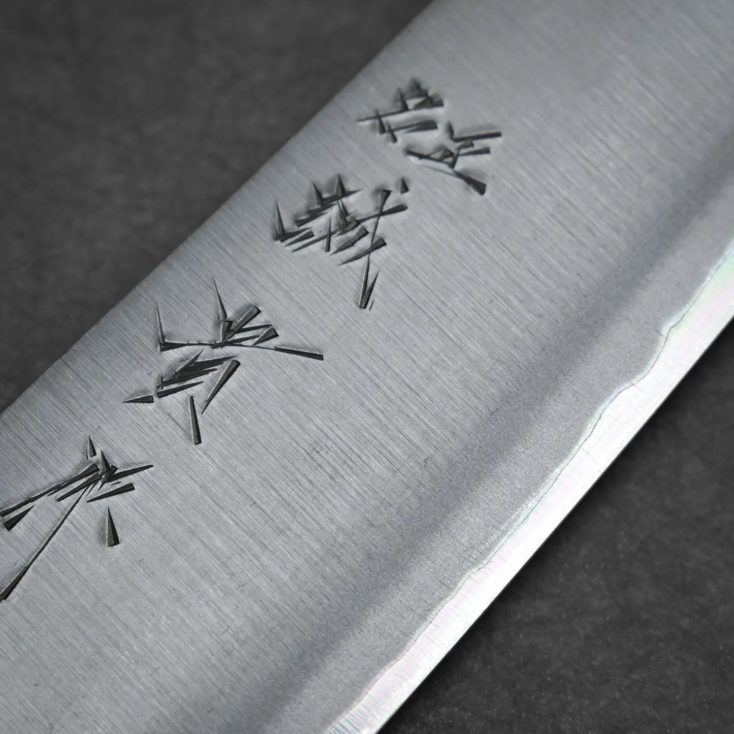 Close-up of a kasumi gyuto blade with Japanese characters engraved near the edge, highlighting the fine texture and sharpness of this workhorse gyuto against a dark background.
