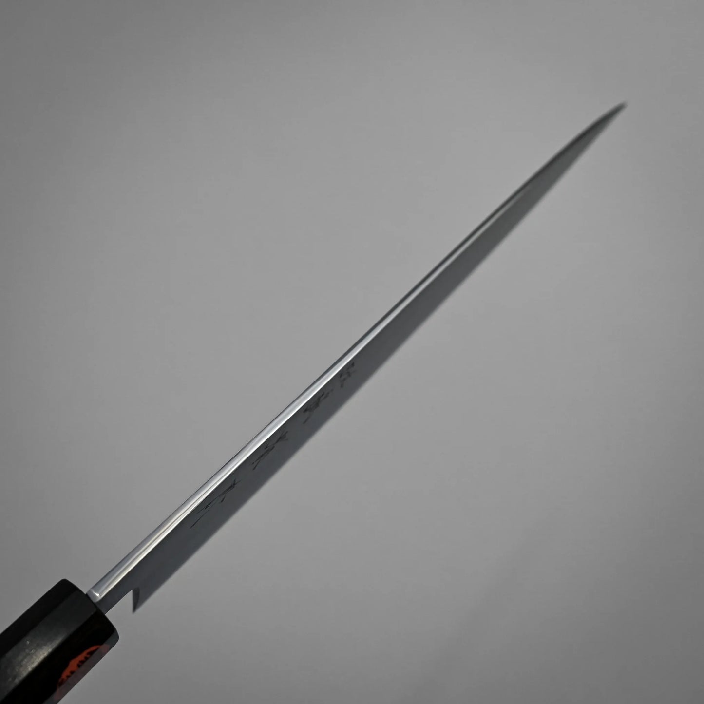 Spine shot of a sharp kasumi gyuto with a black handle, positioned diagonally against a plain gray background. The blade is long, thin, and metallic, casting a slight shadow.