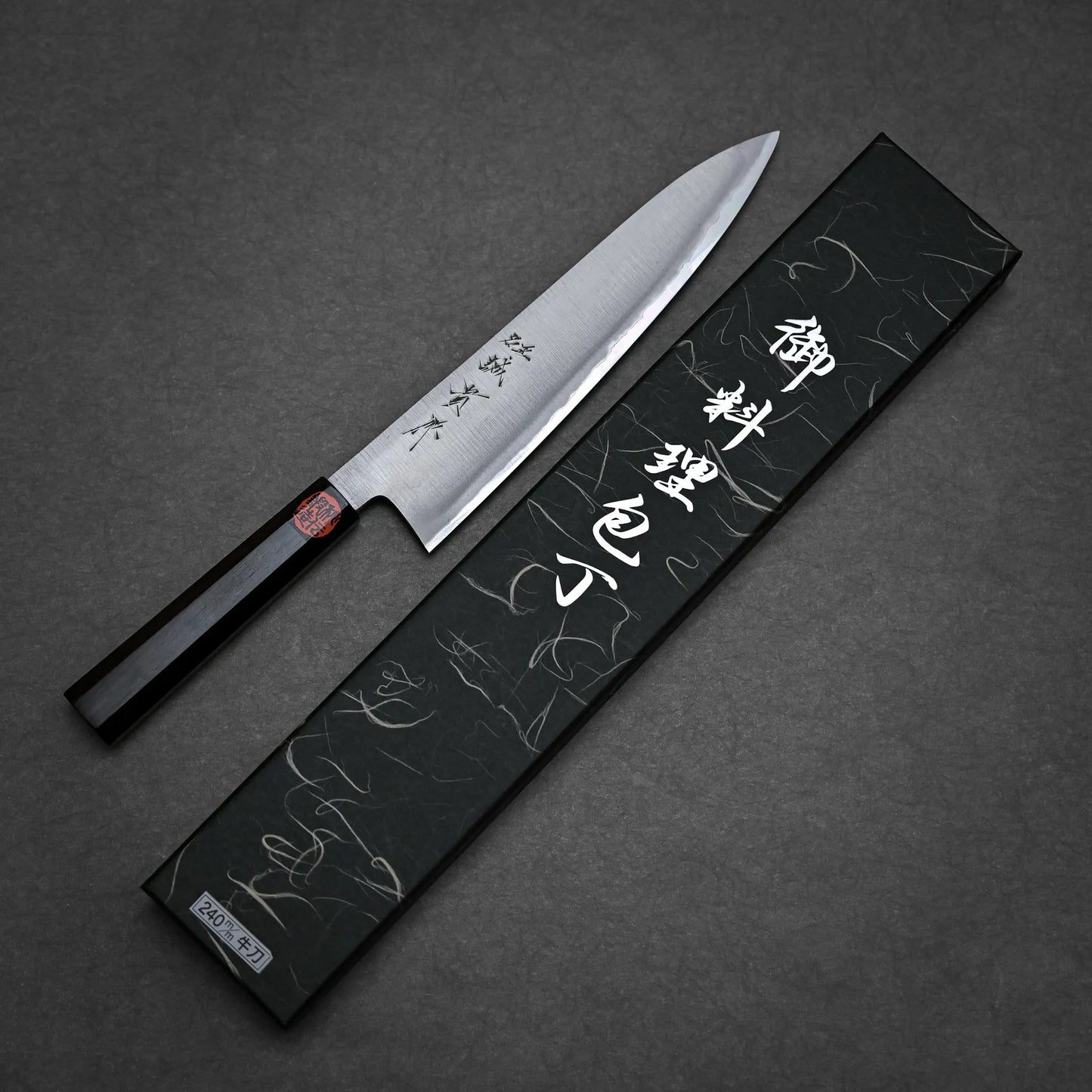 A workhorse gyuto with a dark wooden handle lies beside its decorative black box, which displays silver Japanese characters. Both are set on a dark grey surface.