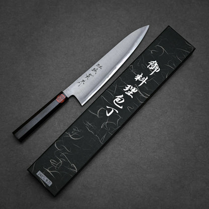 A workhorse gyuto with a dark wooden handle lies beside its decorative black box, which displays silver Japanese characters. Both are set on a dark grey surface.