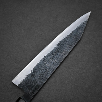 A close-up of a kiritsuke gyuto Japanese chef’s knife with a sharp blade and textured, dark finish near the edge, set against a dark, plain background.