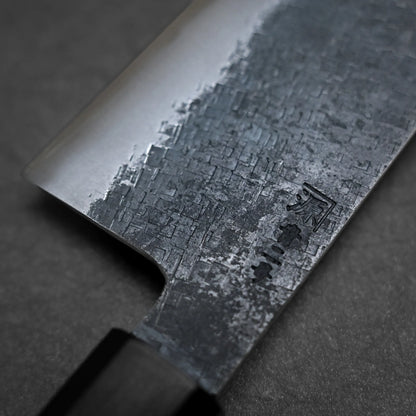 Close-up of a kiritsuke gyuto knife with a textured, patterned blade and engraved kanji characters, set against a dark background. The handle is partially visible.