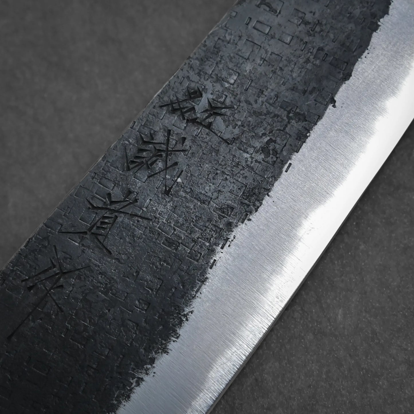 Close-up of a kiritsuke gyuto blade with a textured, dark upper section and a smooth, polished lower edge. Japanese characters are etched into the textured area. The background is a plain dark surface.