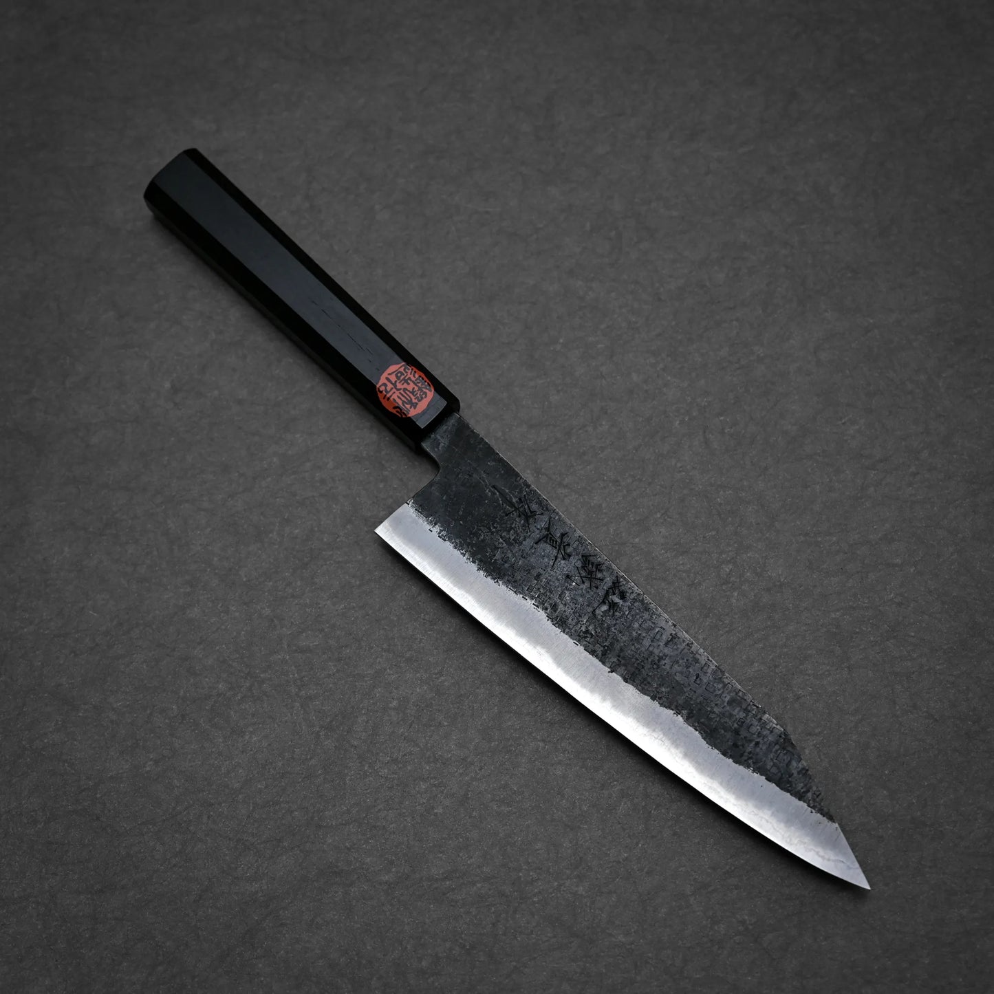 A Japanese-style kurouchi ktip gyuto with a rectangular black handle and a dark, partially rough-textured blade rests on a gray surface. The blade features subtle markings and a red sticker near the handle.