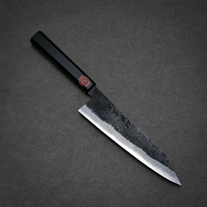 A Japanese-style kurouchi ktip gyuto with a rectangular black handle and a dark, partially rough-textured blade rests on a gray surface. The blade features subtle markings and a red sticker near the handle.