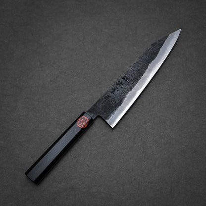 A Japanese-style kurouchi ktip gyuto with a dark, textured blade and a black, octagonal handle is placed on a dark gray surface.