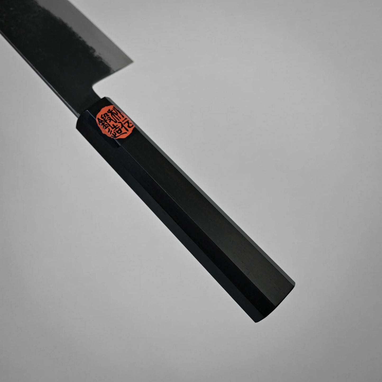 Close-up view of a black, faceted handle on a kurouchi ktip gyuto with a red and black sticker near the blade, photographed against a plain gray background. Only part of the workhorse gyuto's blade and handle are visible.