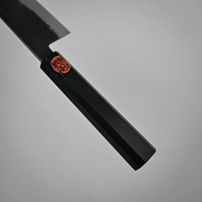 Close-up view of a black, faceted handle on a kurouchi ktip gyuto with a red and black sticker near the blade, photographed against a plain gray background. Only part of the workhorse gyuto's blade and handle are visible.