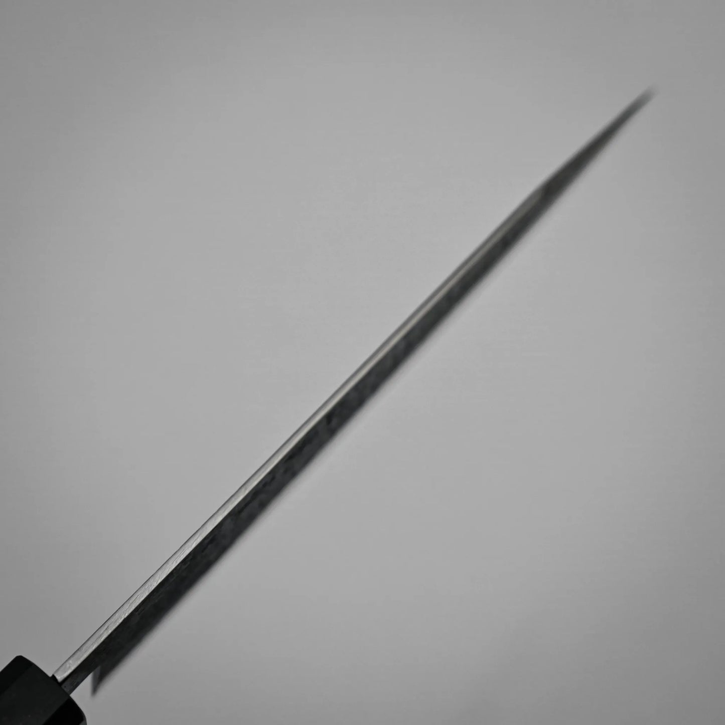 Spine shot of a workhorse gyuto blade photographed from the handle, with the sharp edge extending diagonally across a plain, light background. The blade is in focus near the handle and becomes blurred toward the tip.
