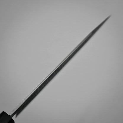 Spine shot of a workhorse gyuto blade photographed from the handle, with the sharp edge extending diagonally across a plain, light background. The blade is in focus near the handle and becomes blurred toward the tip.