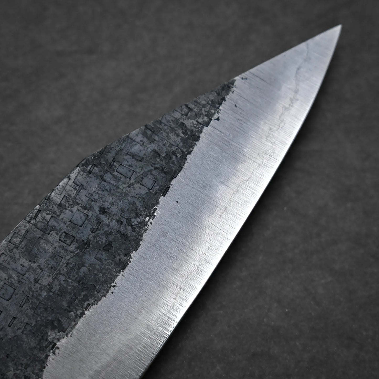 Close-up view of a workhorse gyuto blade, featuring a textured, dark upper section and a smooth, polished lower edge against a plain dark background.