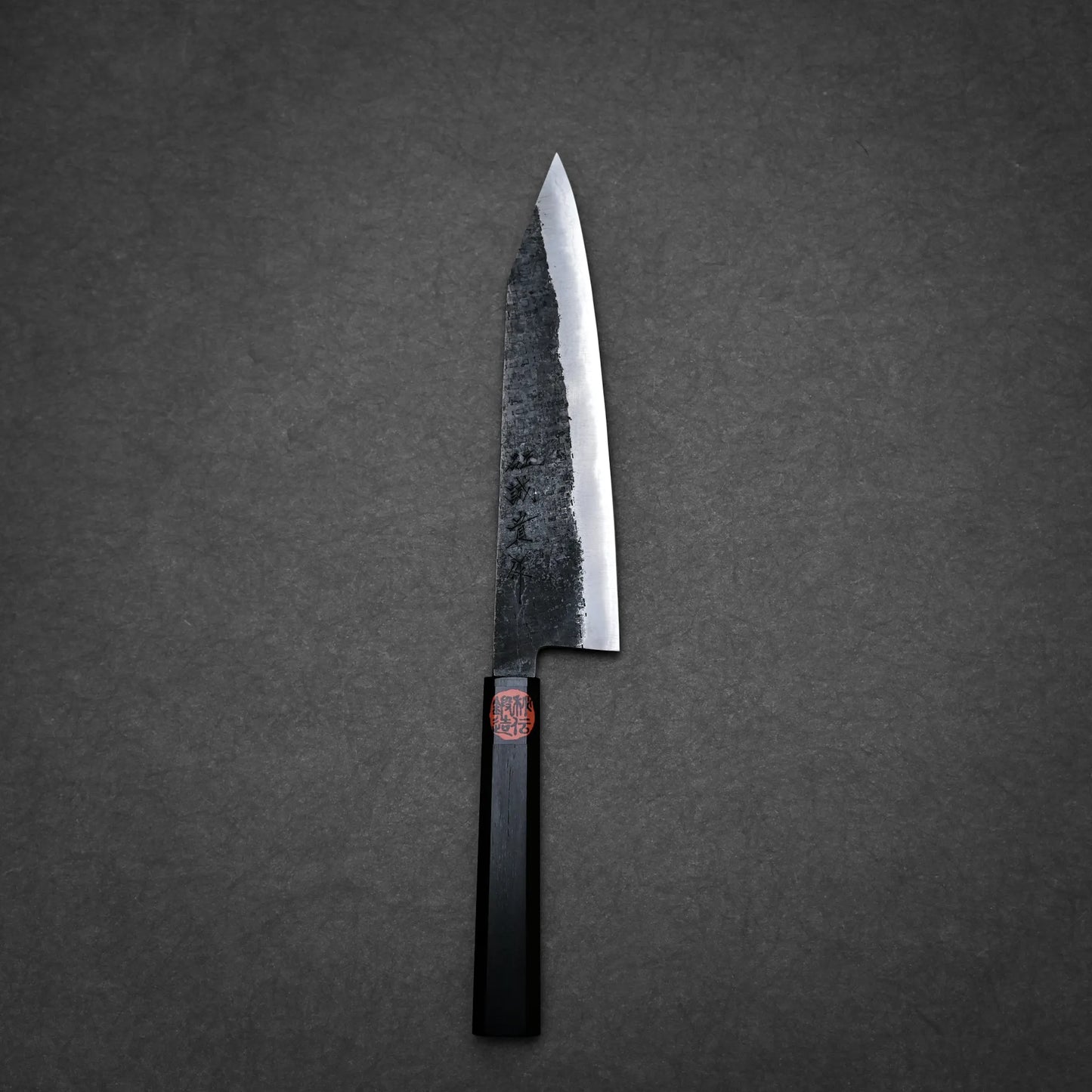 A Japanese-style kurouchi ktip gyuto with a black, rustic, textured blade and a dark, angular handle rests on a dark gray, textured surface.
