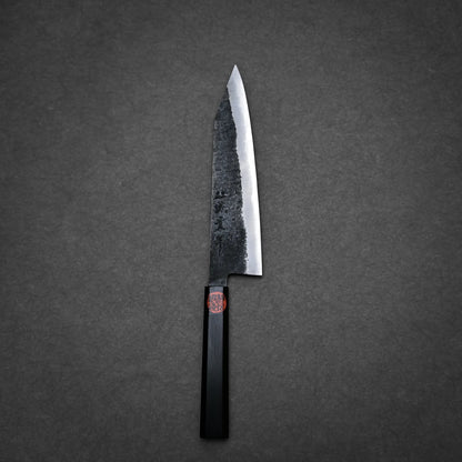A Japanese-style kurouchi ktip gyuto with a black, rustic, textured blade and a dark, angular handle rests on a dark gray, textured surface.