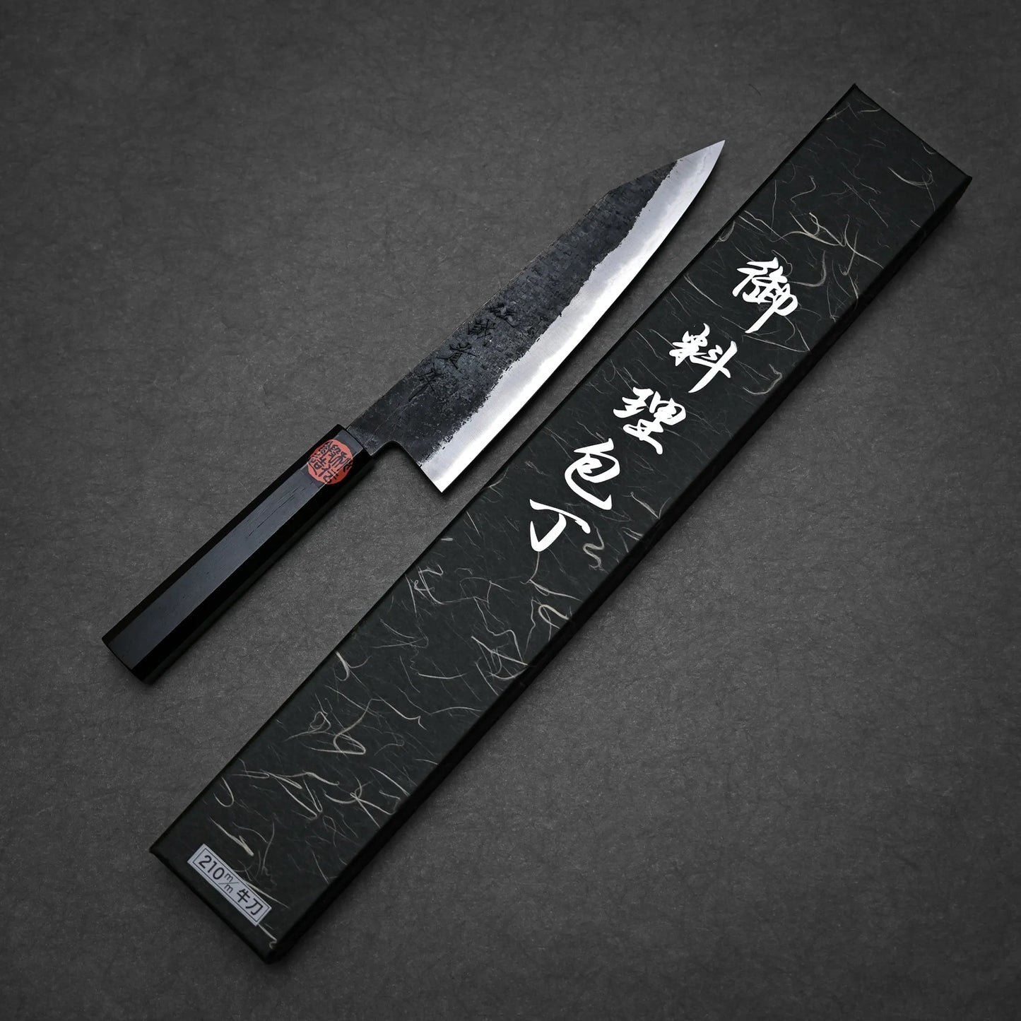 A kurouchi ktip gyuto with a black handle and textured blade rests beside an ornate black box adorned with white Japanese characters, set against a dark background.